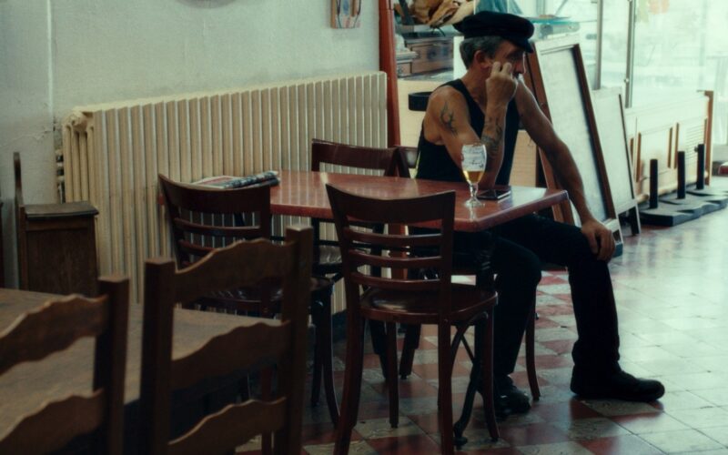 Capture d'écran du film Atlantic Bar, montrant un homme d'une cinquantaine d'années, portant un beret et un marcel noirs, attablé à une table avec une bière
